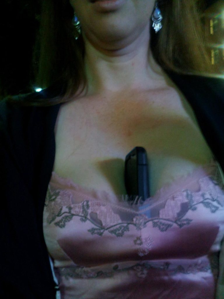 selfie of the front of a woman with a cellphone stuck down her shirt in her cleavage