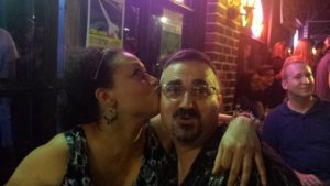 someone makes a face at the camera while another person kisses his cheek dramatically while a third person in the background smiles