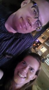 a short person leans on a very tall person for a selfie in an evening city scene