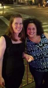 two women smile at the camera with a nighttime city intersection behind them