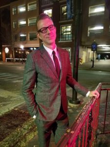 a person dressed elegantly in a suit poses against a railing on a city corner