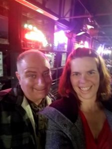 a selfie of two people in coats with bright blurry bar neon signs behind them