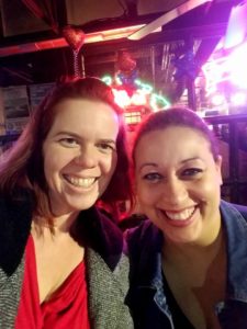 a selfie of two people in coats with bright blurry bar neon signs behind them