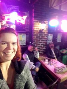 a sideways selfie of a person smiling and a view of two people at a bar table lit with harsh neon behind her