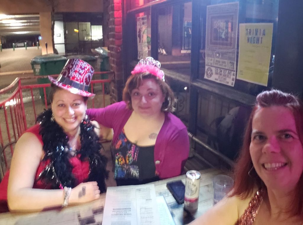 three people at a bar table pose for a selfie wearing new years eve hats and feathers