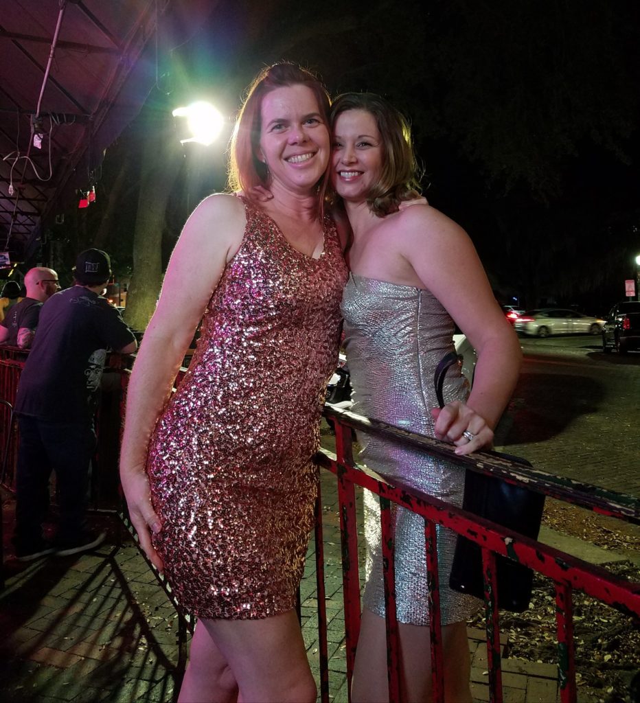 two women in glittery dresses pose for a photo against a railing in an evening city scape