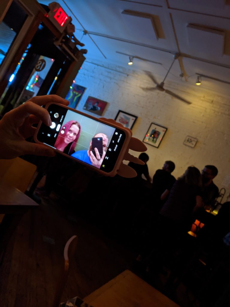 a smart phone is raised in a dark bar to show someone with another phone taking a selfie of two people