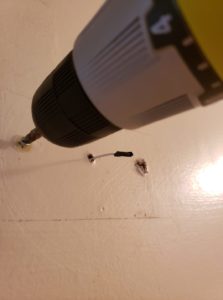 close up of a drill turning a screw in the wall with a hole nearby and a narrow wire protruding