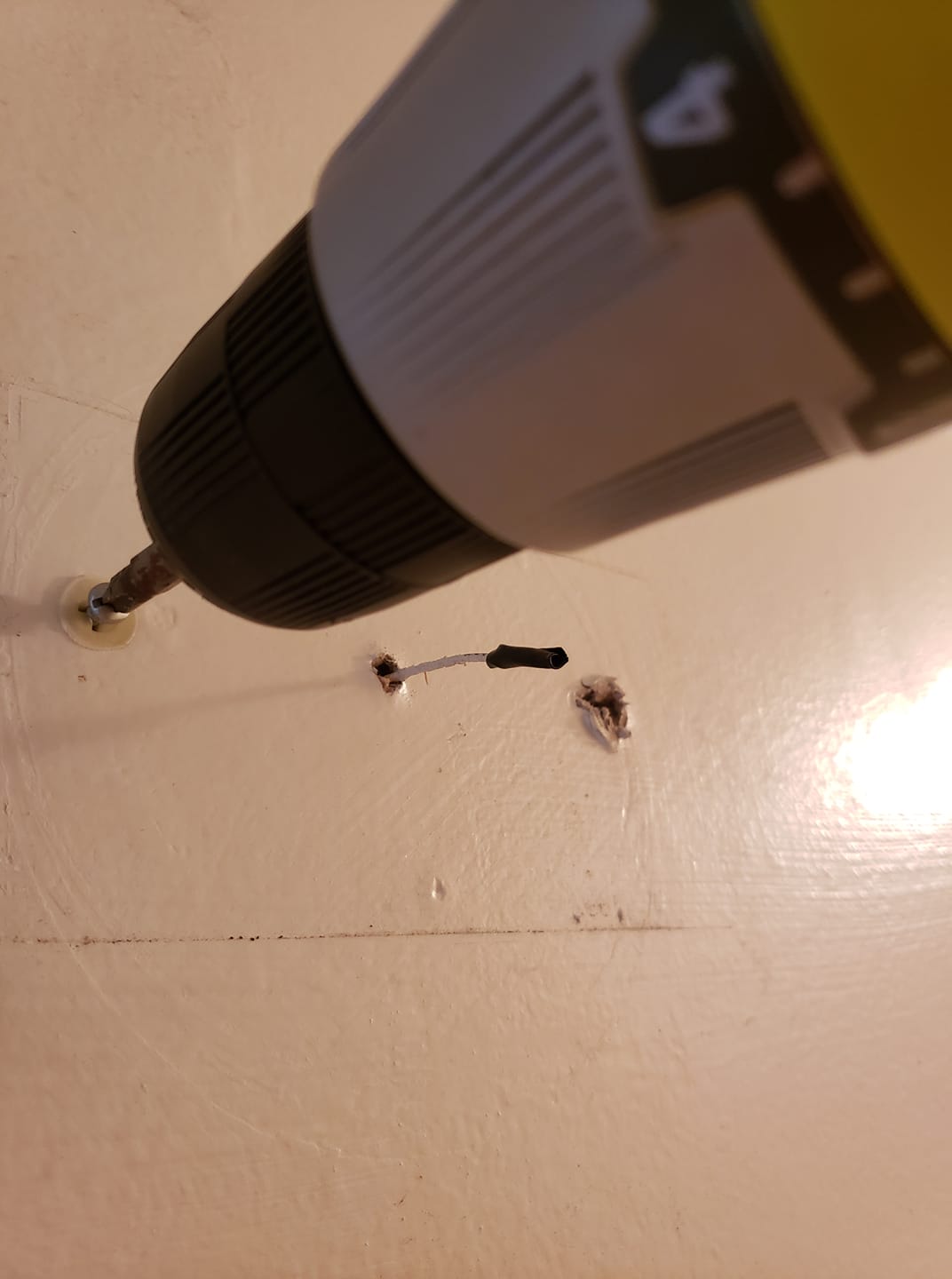 close up of a drill turning a screw in the wall with a hole nearby and a narrow wire protruding