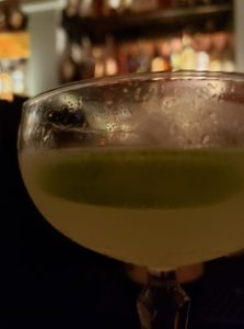 close up of a cocktail glass with cloudy fluid in it and illuminated bar shelves behind it