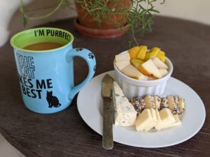 a plat f sliced cheeses and a small bowl of cut fruit next to a colorful mug of coffee with the phrase The Cat Likes Me Best on it