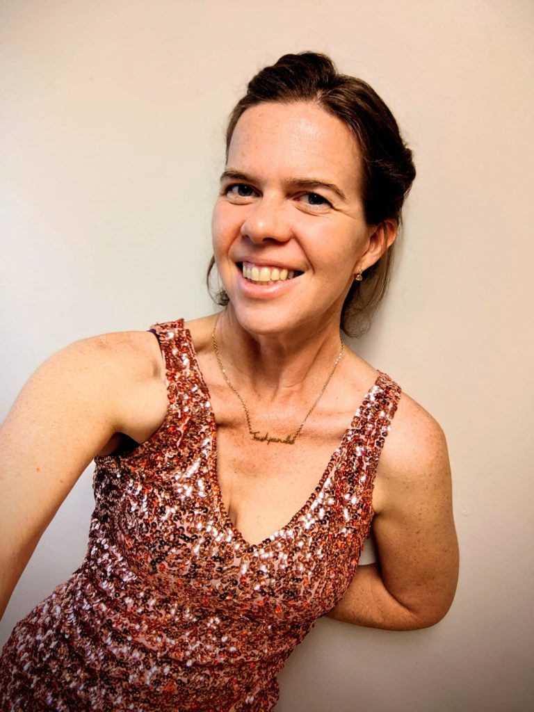 a selfie of a smiling person in a glittery gold dress holding her arm behind herself to lean on the nearby wall