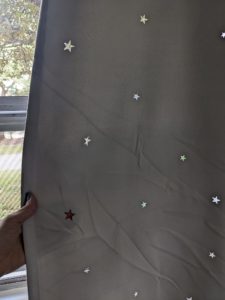 a hand holding slightly wrinkly grey fabric across a window showing the star shaped holes and the light shining through them