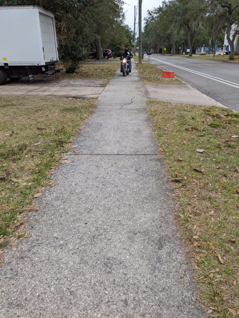 a sidewalk in a residential area with a person on a small motorcycle driving on the sidewalk