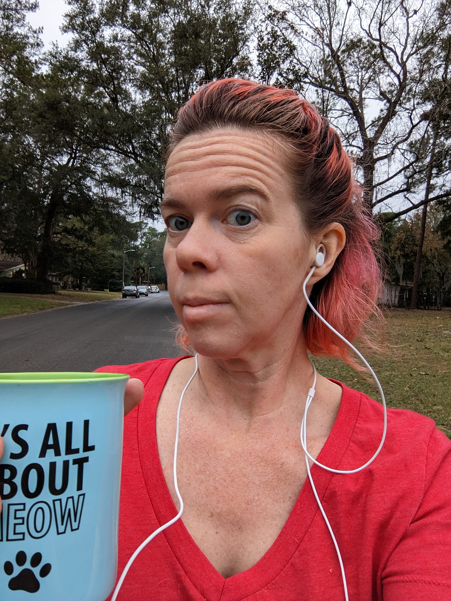 a selfie of a person in a red shirt and earphones holding a coffee mug and walking on a street