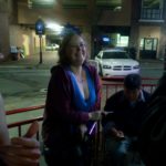 a parson smiles and leans on a railing at night with a city scene and parking garage behind her
