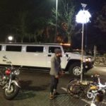 an evening city street with a limo driving past behind a person and their bicycle that has a pole on it with a ball of bright lights