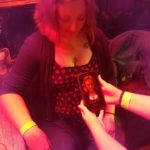 a person sitting on a bar patio is looking down and laughing at hands holding a phone that is taking a picture of her cleavage
