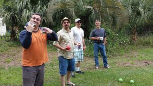 four guys holding balls and beers are standing causally on a lawn together