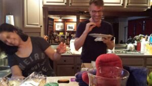 a man holds a plate while eating and a woman is laughing uncontrollably at something while standing in a kitchen