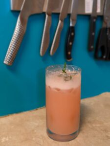 a tall glass with pale pink juice sits on a counter in front of a deeply blue wall under a strip holding a set of knives