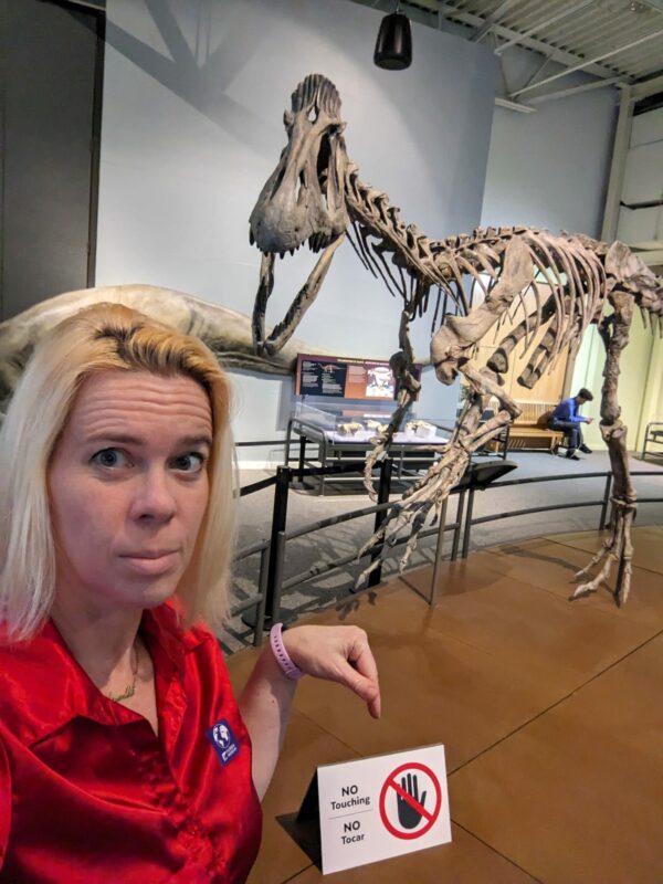 selfie in front of a dinosaur skeleton pointing at a don't tough sign