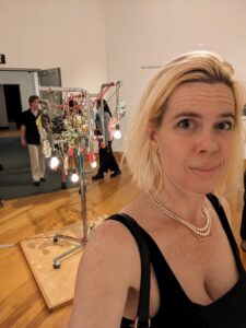 a selfie with a clothes rack looking art sculpture behind me with colorful ornaments and lightbulbs hanging from it