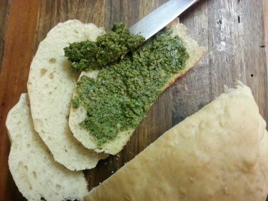 sliced hommade bread is smeared with pesto on a dark cutting board