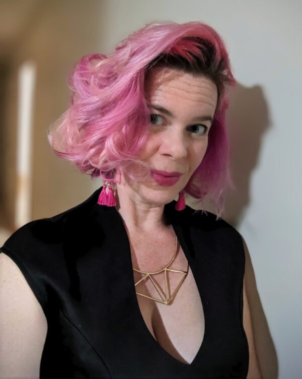 selfie of a lady with vibrant pink hair wearing a black dress and a stylized gold metal necklace