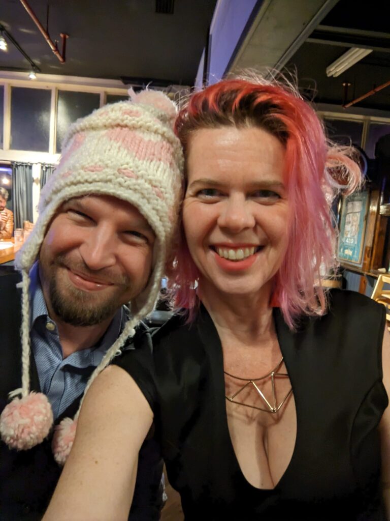 selfie of a woman with pink hair and a black dress with a man wearing a knitted bobble hat with ear flaps with dangling pompoms in a bar of some kind