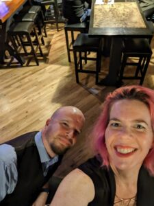 selfie of a woman with pink hair and a black dress with a man in a suit vest with a shiny blond pub floor below them