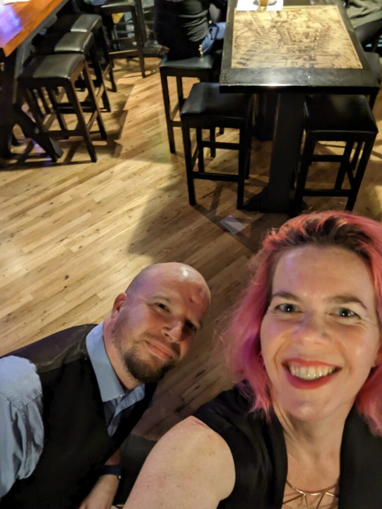 selfie of a woman with pink hair and a black dress with a man in a suit vest with a shiny blond pub floor below them