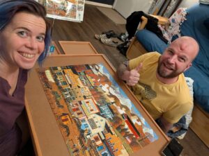two people smiling up at the camera across a short table with a completed puzzle of a vintage painting between them