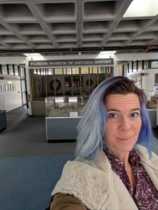 selfie of a woman with blue hair and a coat in an office lobby that looks closed and a sign for a museum