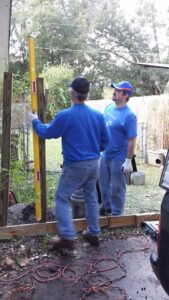 two people are holding wooden beams and a long level in a yard with tools at their feet