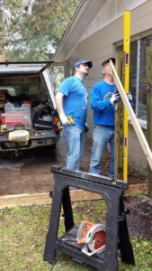 two people are holding wooden beams and a long level in a yard with tools at their feet and a truck behind them