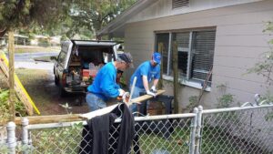 two men are sawing a long piece of wood in a yard with tools around them and a small work truck behind them
