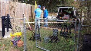 two people are holding wooden beams and a long level in a yard with tools at their feet and a truck behind them