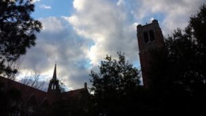 there is some kind of bell tower and spikey buildings like an old city or college and some trees silhouette with a bright cloudy sky in the background