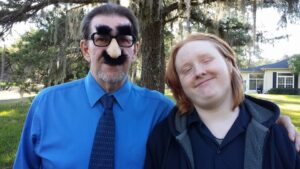 two people smiling at the camera with one wearing novelty glasses with a fake nose and mustache