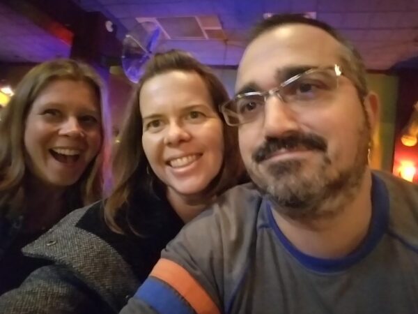 a selfie of three people leaning close with colorful lighting behind them