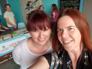 two people with red hair taking a selfie with a table behind with a banner for vegfest