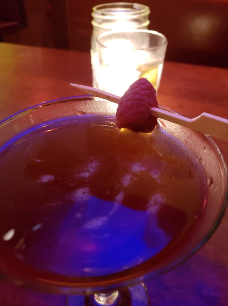 a cocktail with a raspberry on a stick is lit by candles in the back
