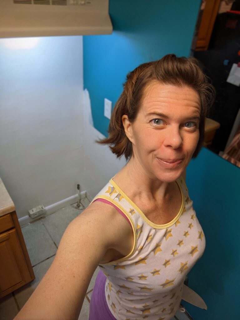 a selfie of a person looking happy with what looks like an empty space in a kitchen cabinetry like a stove or fridge was moved