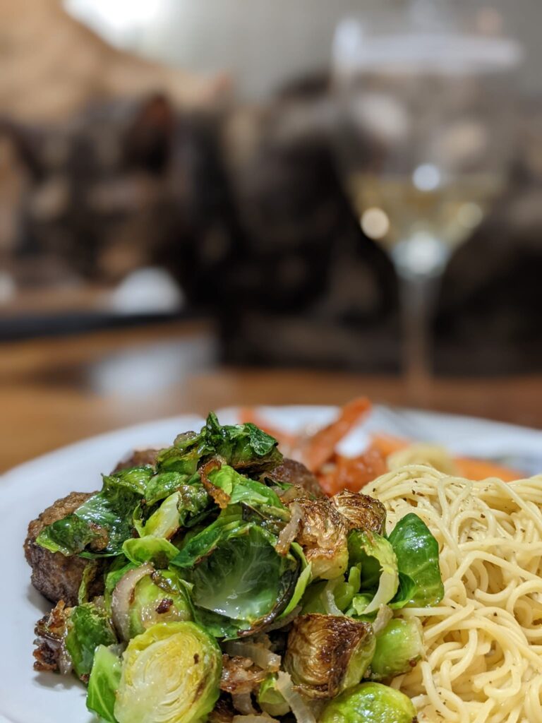 a plate of pasta and brussels sprouts with maybe a cat and a glass of wine blurry in the distance