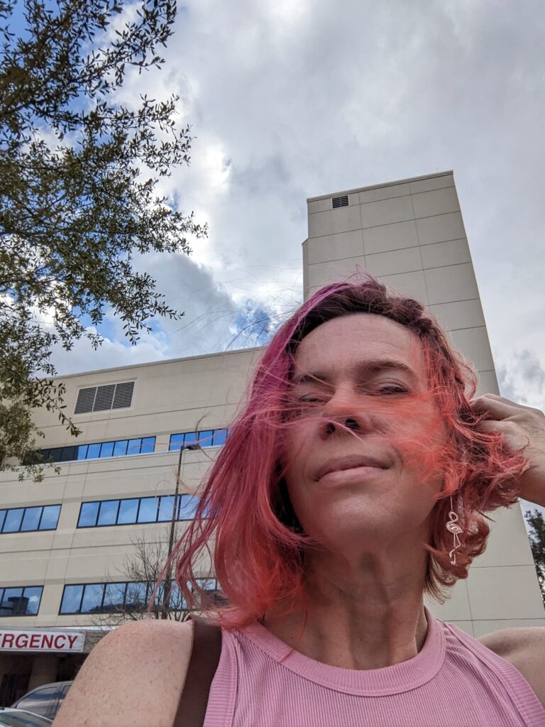 selfie of a person with wind blowing their hair across their face and a hospital building behind them
