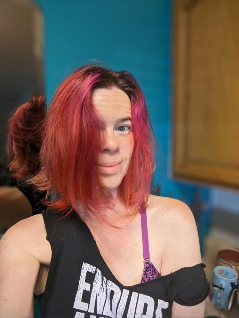 selfie of a person with red and purple hair an a vibrant teal blue wall behind her