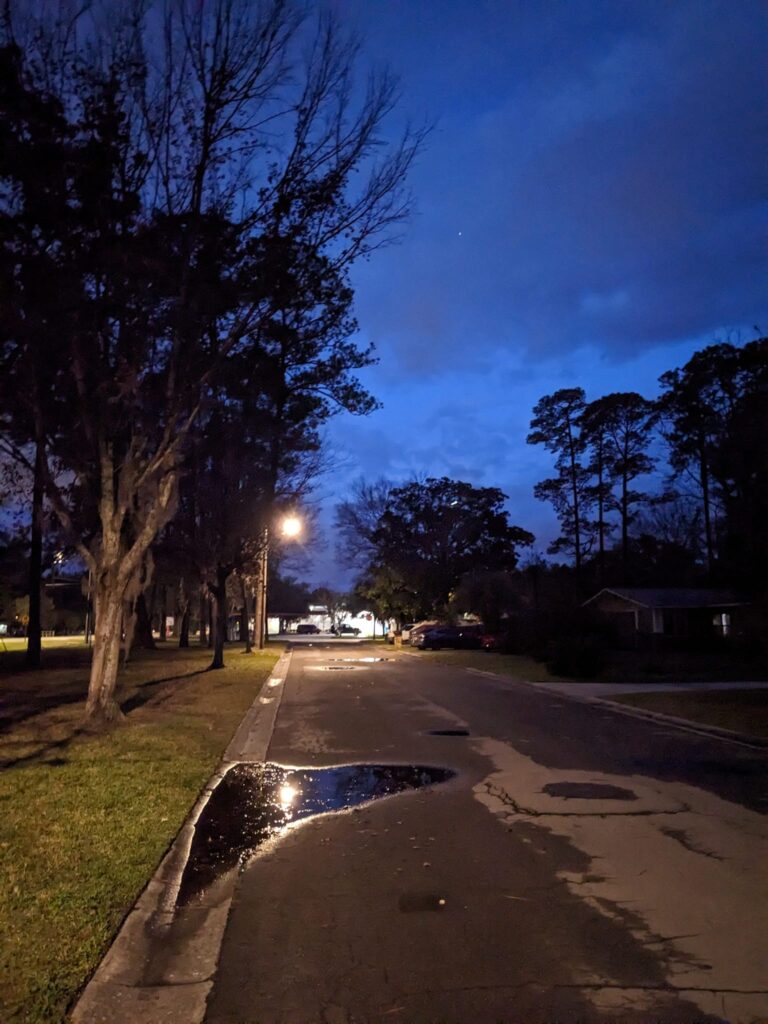 a dark neighborhood street has a street light reflecting on a puddle and a deeply blue cloudy night sky above