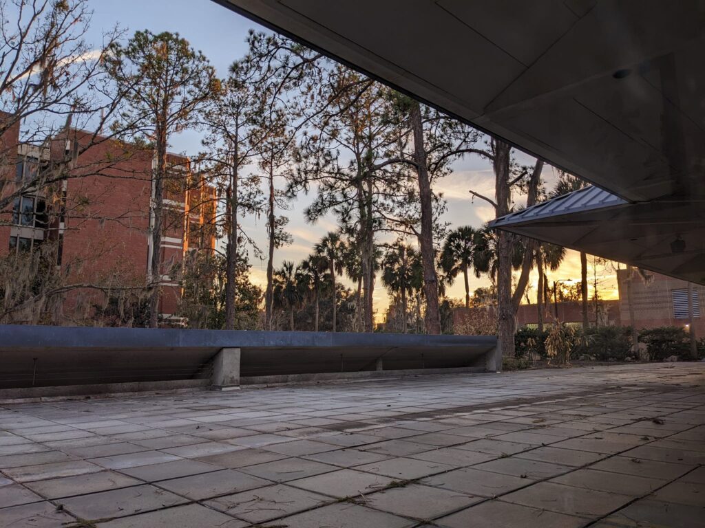 a wide patio surrounded by buildings and a roof overhand has a glowing sunlight in the distance behind a row of trees