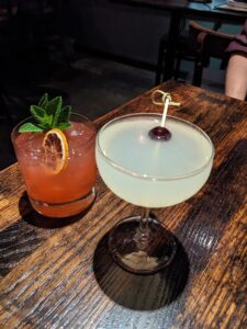 two cocktails on a wooden bar, one short with citrus and mint and one in a champagne style glass is milky white with a cherry